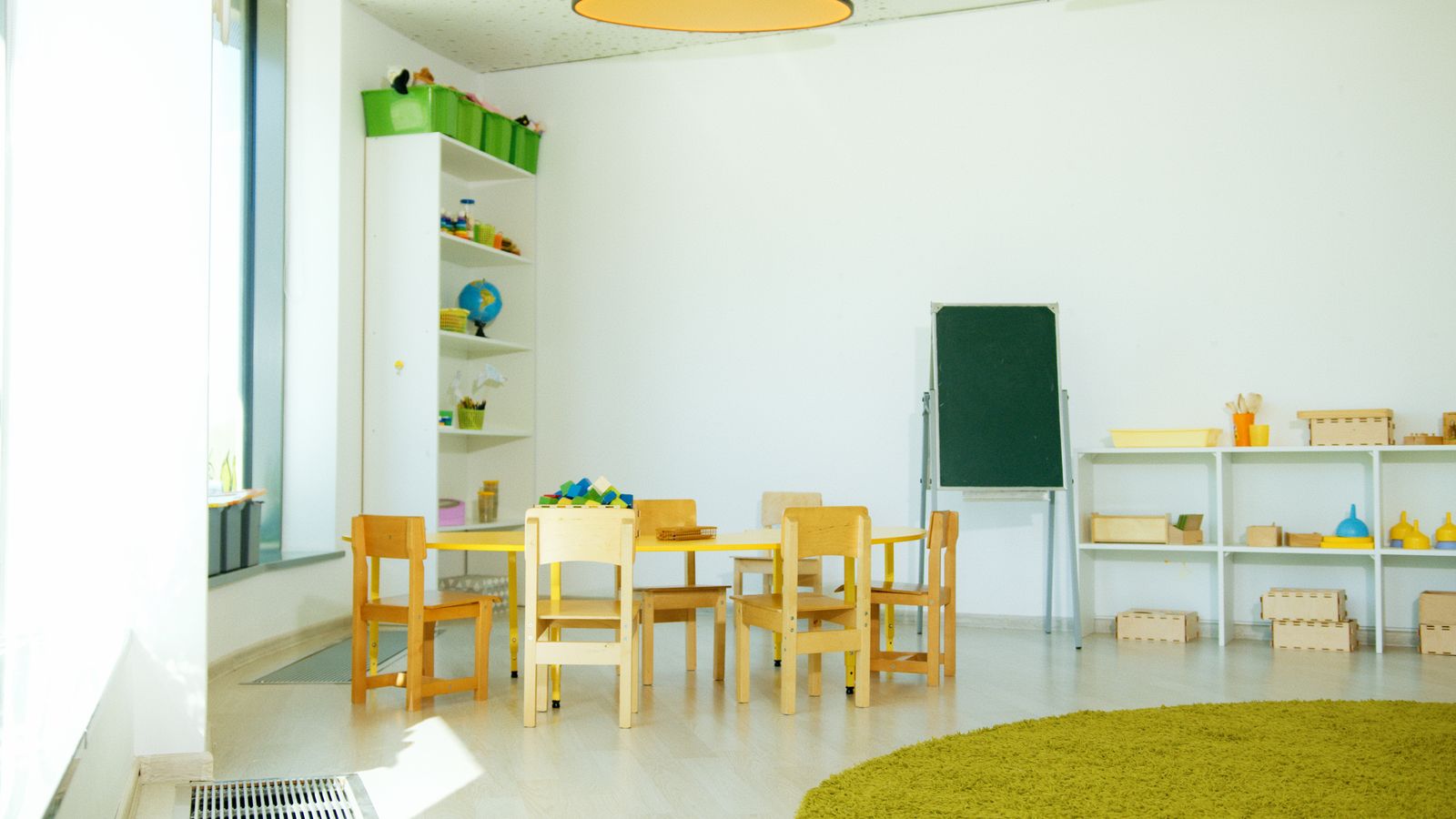 A bright early-childhood classroom with child-sized wooden tables and chairs, learning materials on open shelves, and a round rug.